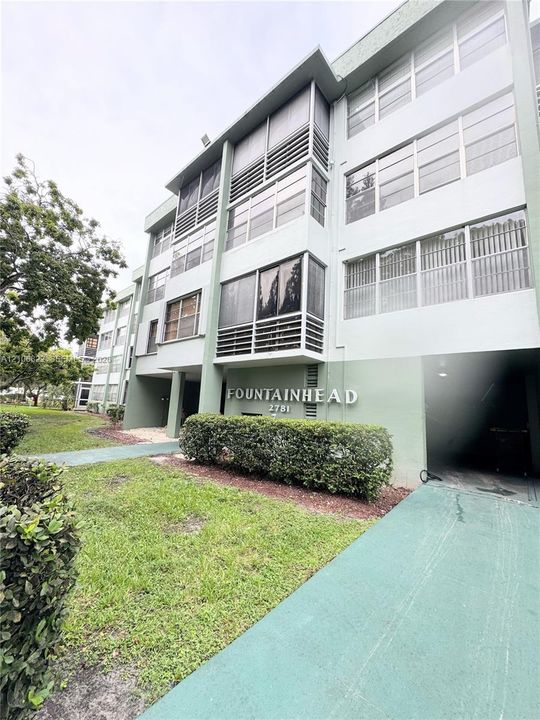 Multi-story building exterior featuring a structured concrete facade, enclosed balconies with louvered screens, and a covered ground-level entryway