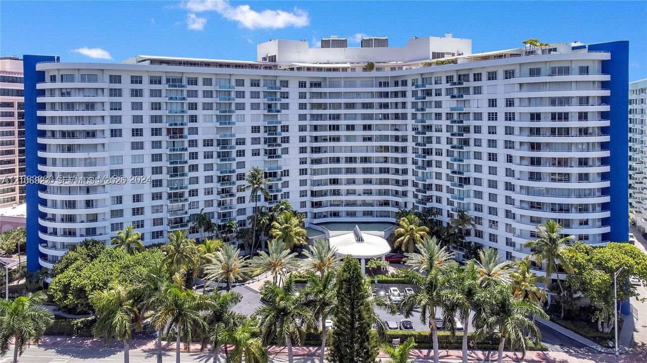 Mid-rise residential building exterior featuring curved balconies, a covered circular drop-off area, blue architectural accents, and mature palm landscaping