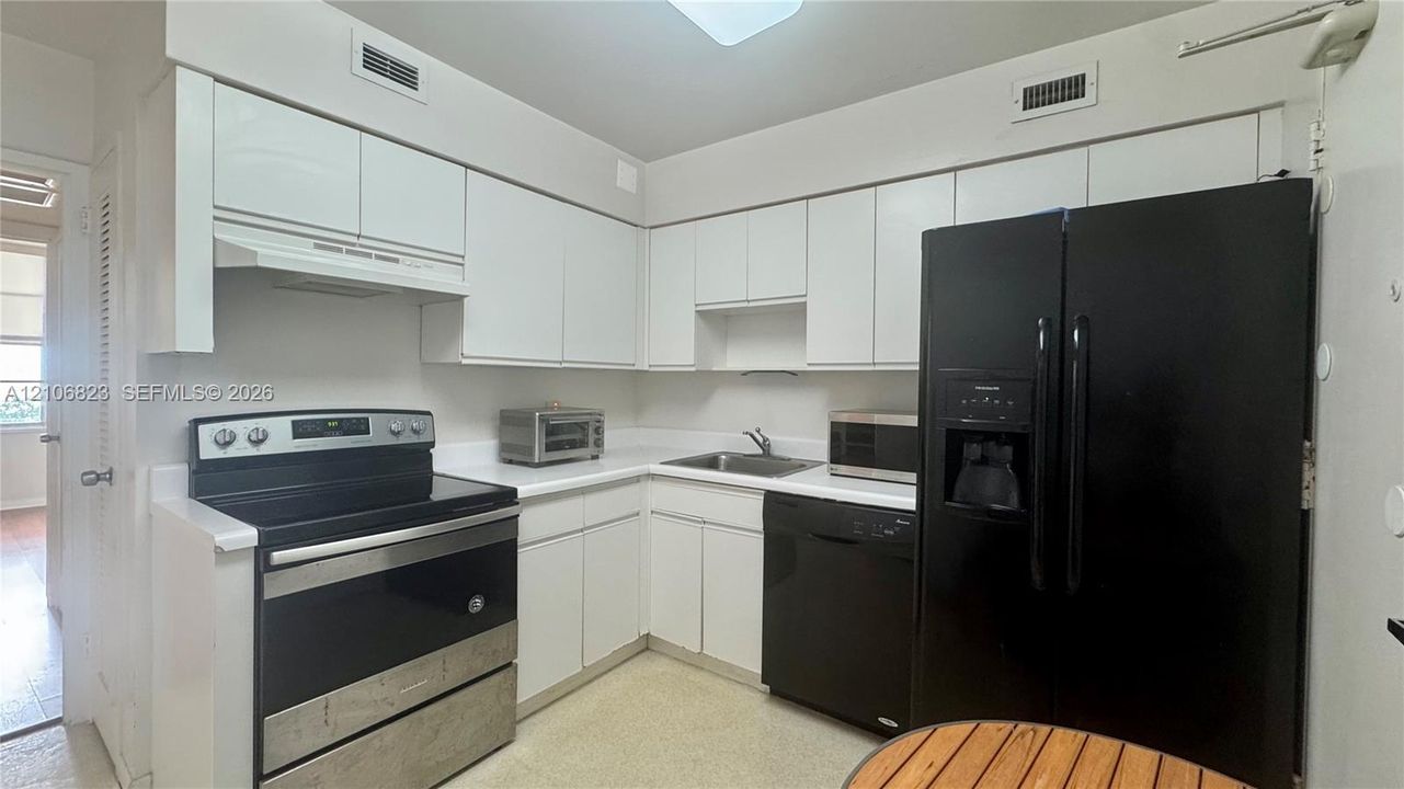 Kitchen featuring white flat-panel cabinetry, solid-surface countertops, integrated range hood, and black appliance suite including a side-by-side refrigerator, dishwasher, and electric range