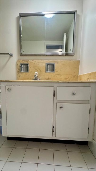 White vanity cabinet with chrome hardware, natural stone countertop with matching backsplash, dual inset soap dishes, and a framed mirror