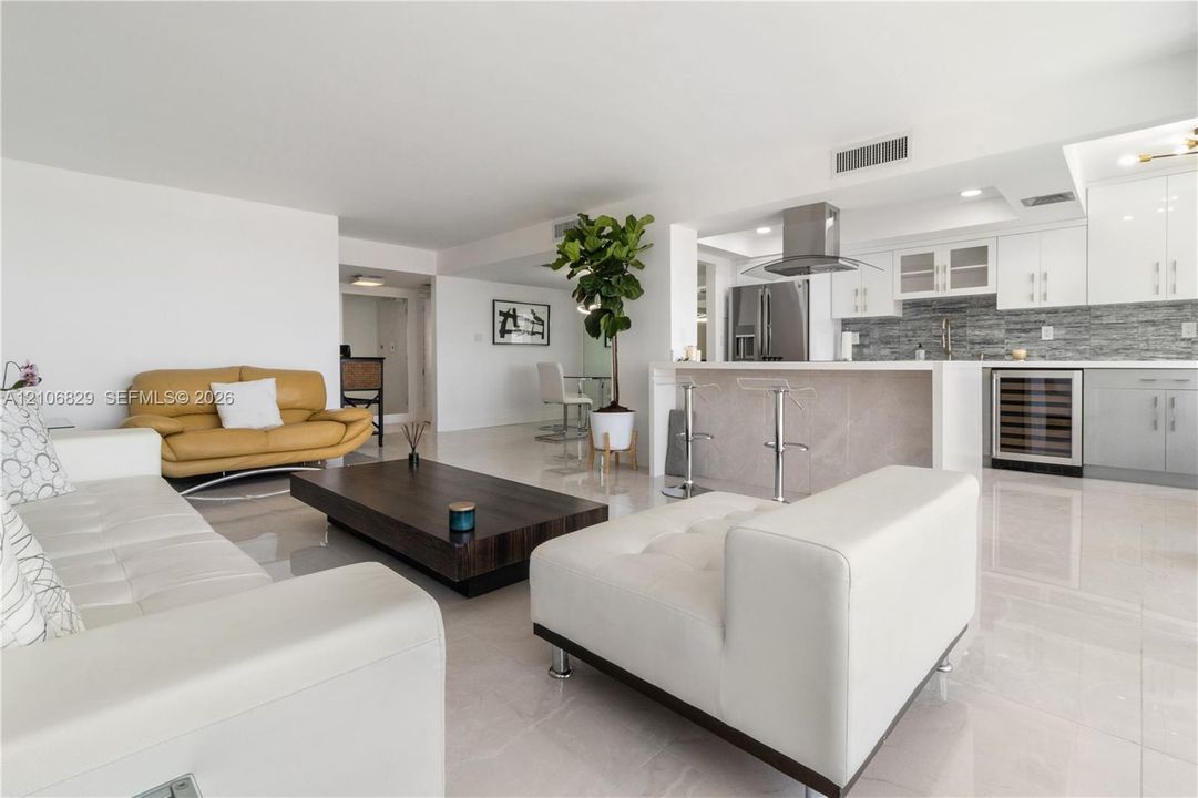 Open-concept living space featuring high-gloss tile flooring, white cabinetry, integrated stainless steel appliances, a kitchen island with a stone-finish facade, and a built-in wine refrigerator