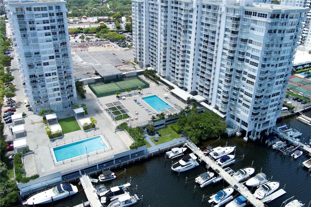 Aerial perspective of a multi-story residential complex featuring two swimming pools, a tennis court, landscaped green space, and private deep-water dockage