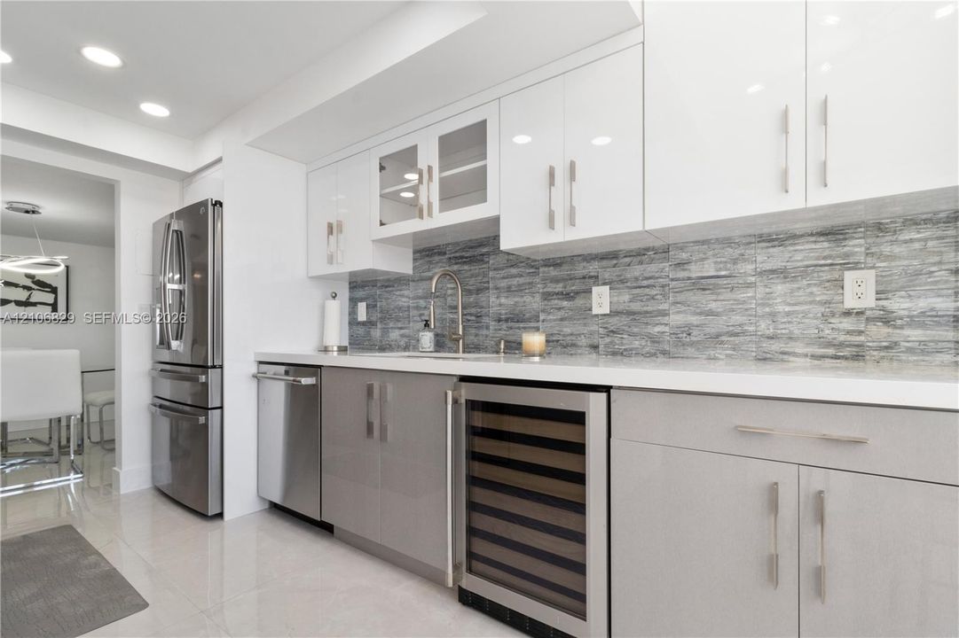Modern kitchen featuring high-gloss white upper cabinetry, grey base cabinetry, integrated wine refrigerator, stainless steel appliances, and a horizontal-patterned stone backsplash