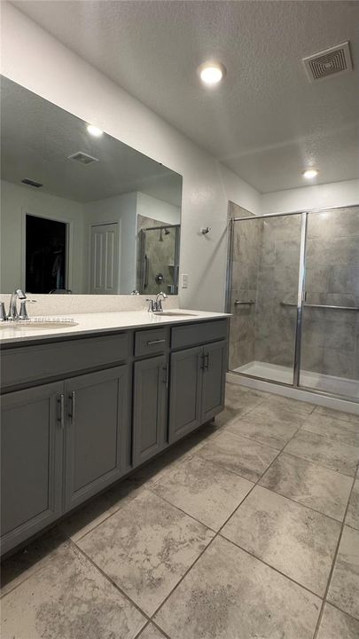 Dual-sink vanity with gray shaker-style cabinetry, white countertop, large wall-to-wall mirror, and glass-enclosed shower with stone-finish tile surround