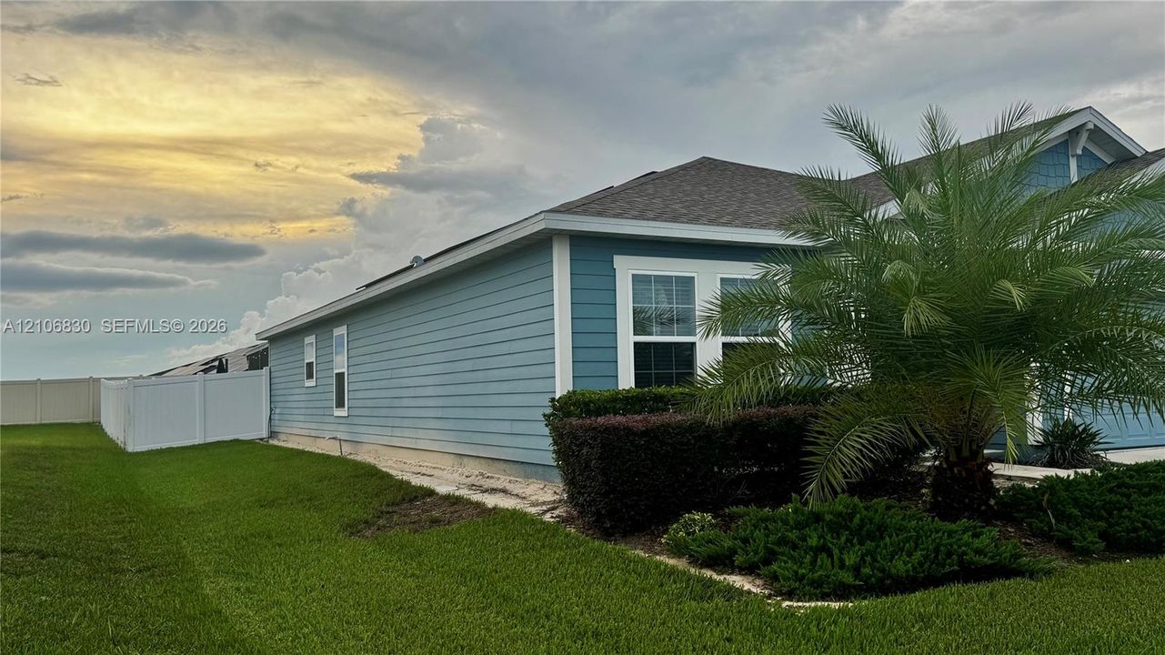 Exterior facade featuring horizontal lap siding, a pitched shingled roof, white window trim, mature palm landscaping, and a vinyl privacy fence