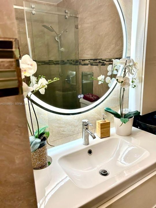 Wall-mounted circular LED mirror, white integrated sink with chrome single-handle faucet, and glass-enclosed shower with tiled walls and decorative mosaic accents