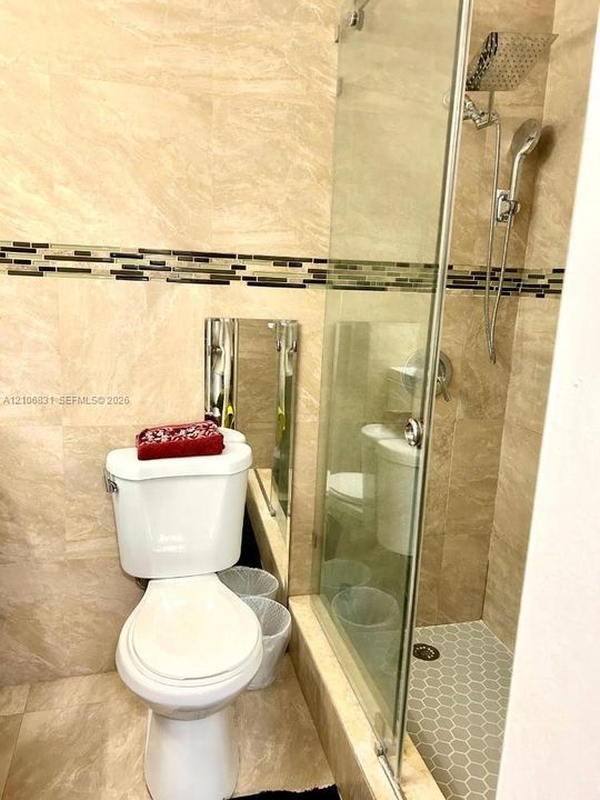 Bathroom featuring floor-to-ceiling stone-finish tile, glass-enclosed shower with a rainfall showerhead and handheld sprayer, and an integrated glass mosaic accent border