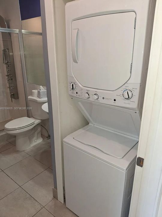 White stacked washer and dryer unit, tiled flooring, porcelain toilet, and a walk-in shower enclosure featuring a multi-jet rainfall shower system