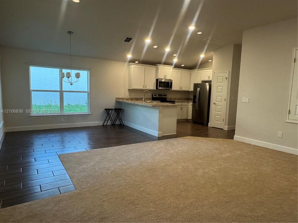 Open-concept living space featuring wood-finish tile flooring, neutral carpeting, white shaker-style cabinetry, a breakfast bar with stone-finish countertops, and stainless steel appliances