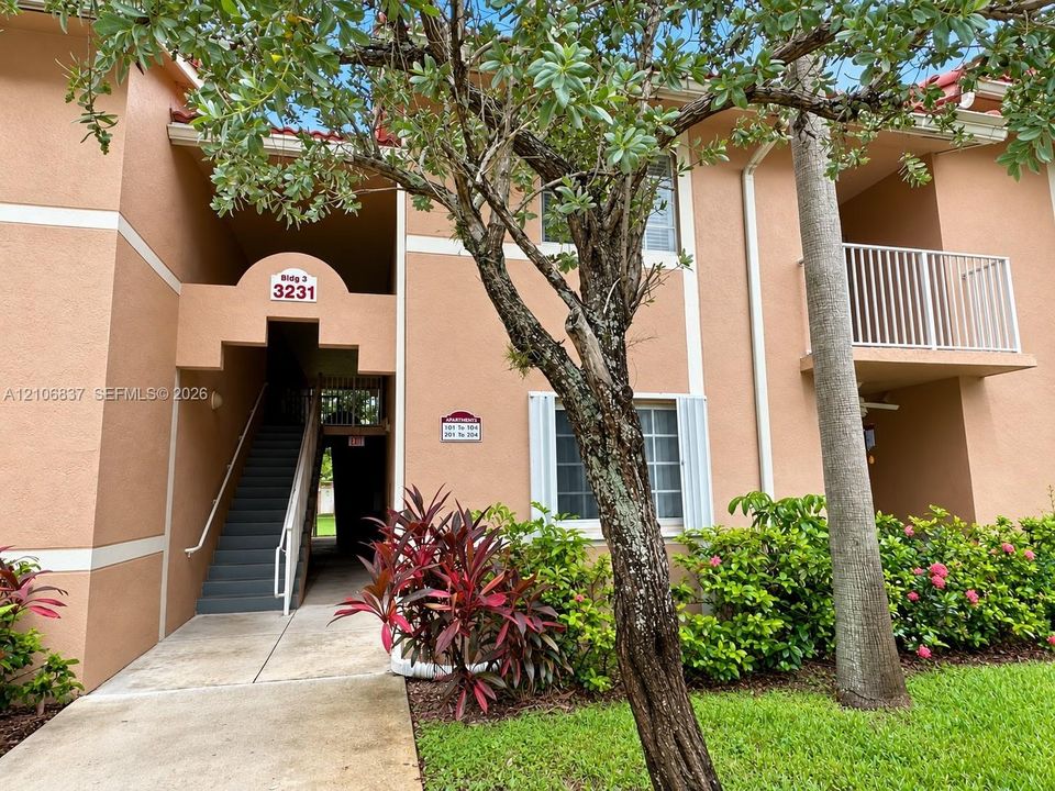 Stucco exterior building featuring a covered second-story balcony with metal railing, concrete walkway, and landscaped garden beds