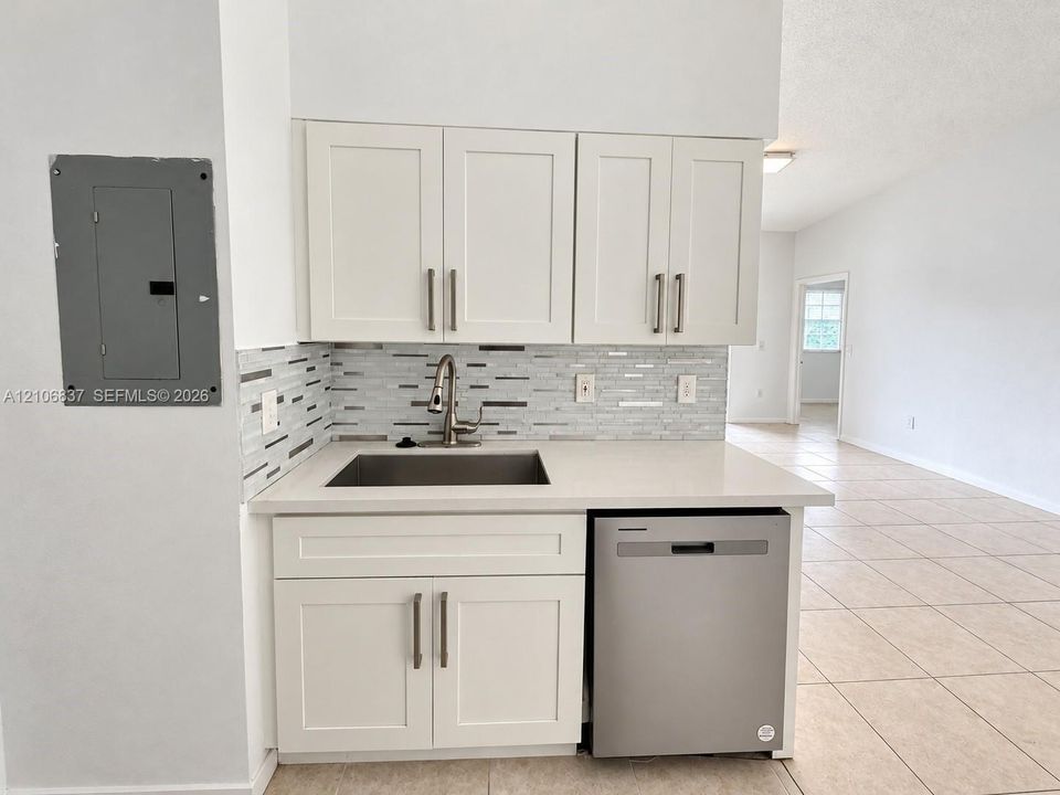 Kitchenette featuring white cabinetry with brushed metal hardware, solid-surface countertops, a stainless steel undermount sink, a mosaic tile backsplash, and an integrated stainless steel dishwasher
