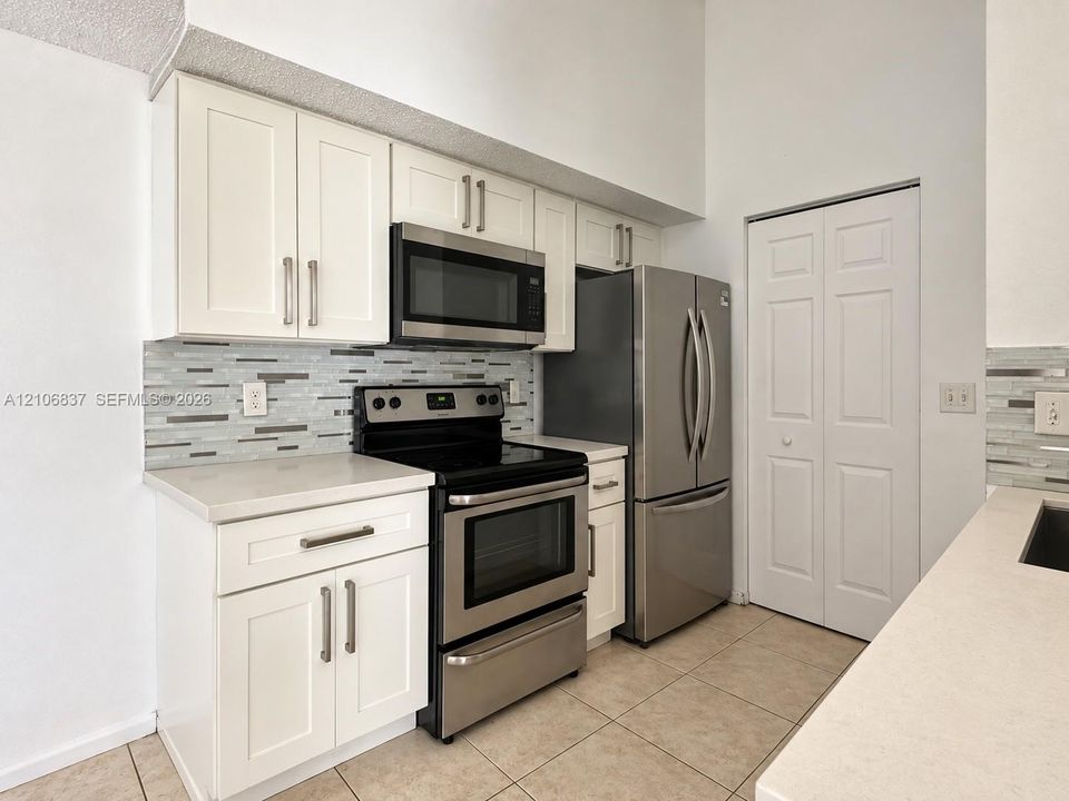 Kitchen featuring white shaker-style cabinetry, quartz countertops, multi-toned linear tile backsplash, stainless steel appliances, and tile flooring