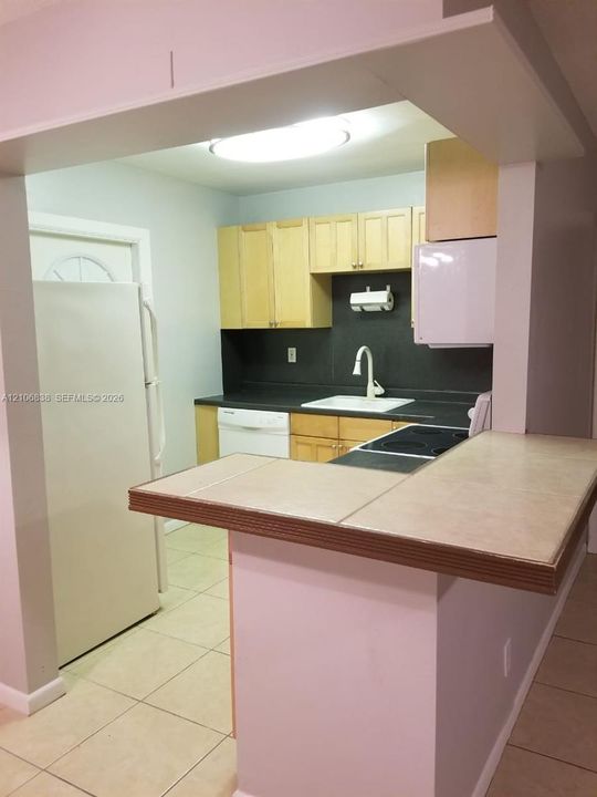 Kitchen featuring light wood cabinetry, a tile-topped breakfast bar, dark countertops, a white top-mount sink with a high-arch faucet, and ceramic tile flooring