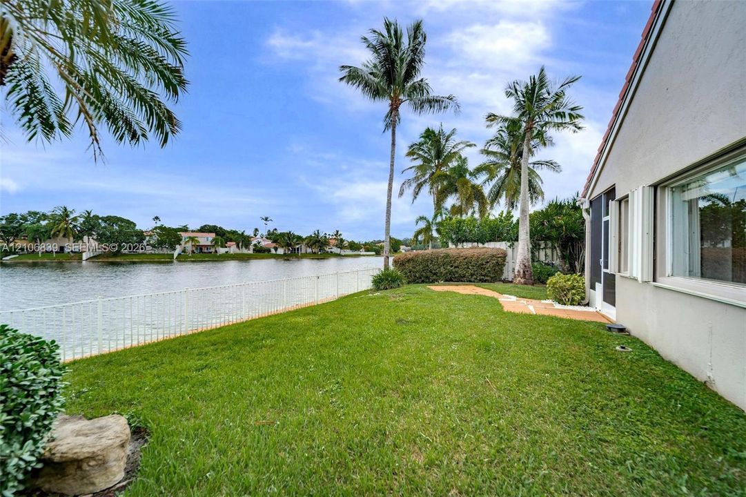 Waterfront exterior featuring a grass lawn, metal fencing, stone-finish paving, and mature palm trees