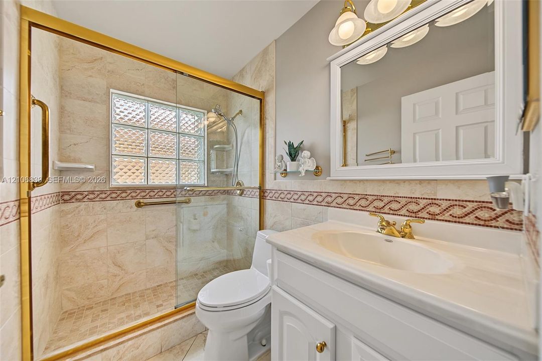 Bathroom featuring a walk-in shower with gold-finish trim, decorative tile accents, a stone-surround vanity with an integrated basin, and a glass-paneled privacy window
