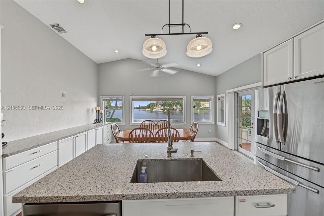 Kitchen island featuring a stone countertop with an undermount stainless steel sink, vaulted ceilings, white shaker-style cabinetry, stainless steel refrigerator, and recessed lighting