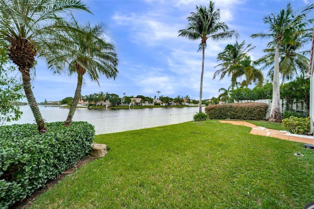Waterfront backyard featuring a manicured lawn, palm trees, mature hedges, and a white perimeter fence