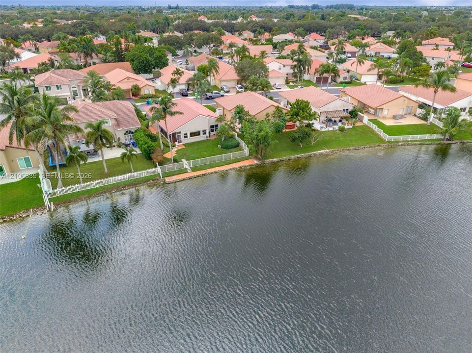 Waterfront residential setting featuring a large body of water, manicured lawns, white vinyl fencing, and palm trees