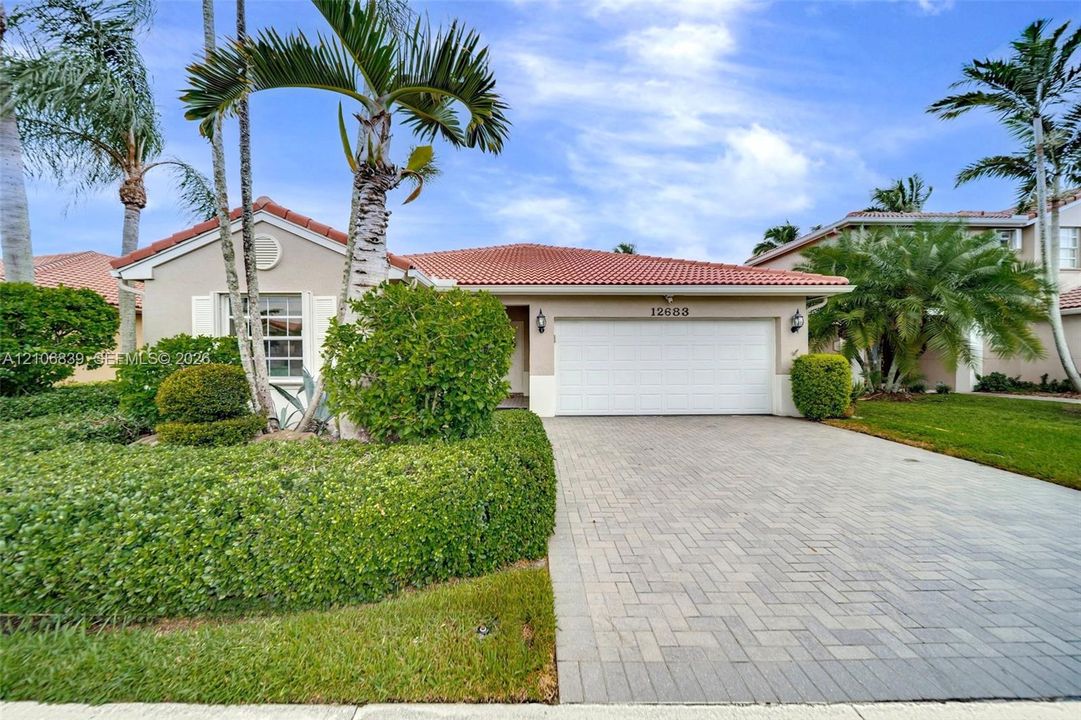 Single-story residence featuring a terracotta-tiled roof, paver driveway, attached two-car garage, and manicured landscaping with mature palm trees