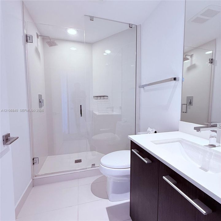 Walk-in shower with frameless glass enclosure, dark-finish cabinetry, solid-surface vanity countertop with integrated sink, polished chrome faucet, and rectangular floor tiling