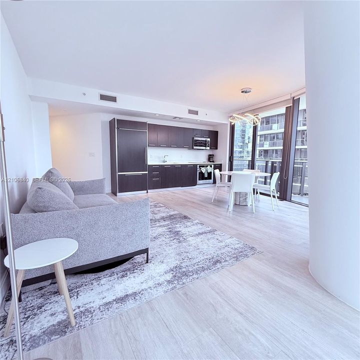Open-concept living space featuring wood-finish flooring, dark cabinetry, stainless steel appliances, floor-to-ceiling glass, and a modern sculptural pendant light fixture