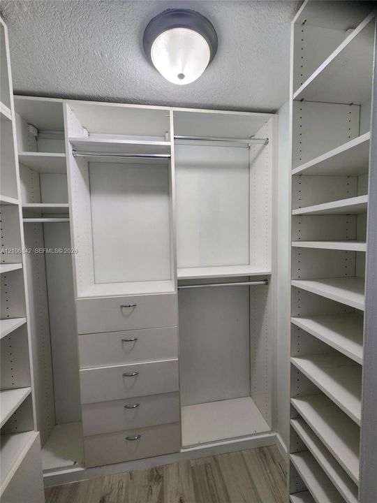 Customized storage closet featuring white built-in shelving, integrated hanging rods, a five-drawer dresser unit, and wood-finish flooring