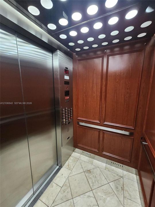 Elevator interior featuring custom wood-panel walls, stone tile flooring, stainless steel sliding doors, a linear metal handrail, and a recessed ceiling panel with circular accent lighting