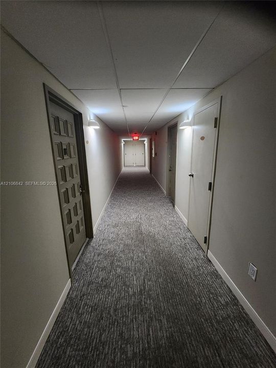 Interior hallway featuring neutral walls, grey patterned carpeting, textured drop ceiling tiles, and wall-mounted sconce lighting