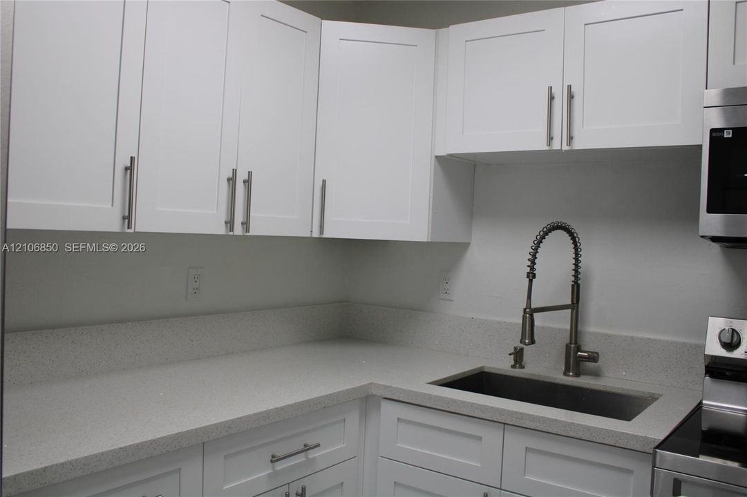 White Shaker-style cabinetry with polished metal hardware, light-toned solid surface countertops, a stainless steel undermount sink, and a high-arch pull-down faucet