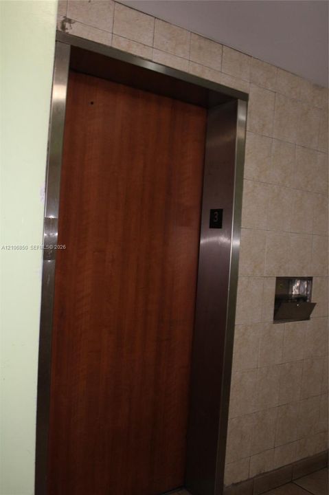 Elevator featuring a wood-finish door, stainless steel frame, and neutral-toned wall tiling
