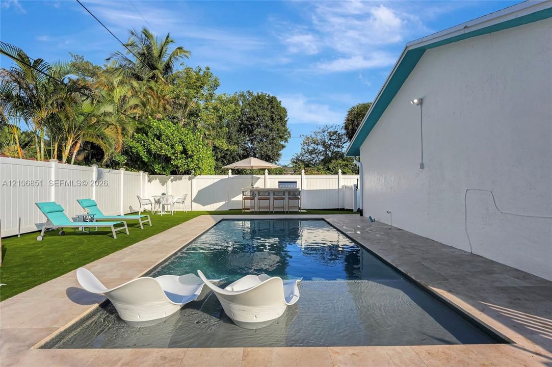 In-ground swimming pool with shallow tanning ledge, complemented by light-toned stone paver decking and synthetic turf lawn, all enclosed by white vinyl privacy fencing