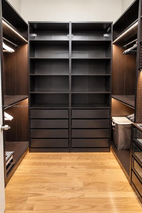 Custom walk-in closet featuring dark-finish built-in cabinetry, extensive open shelving, integrated drawer units, recessed LED lighting, and wood-finish flooring
