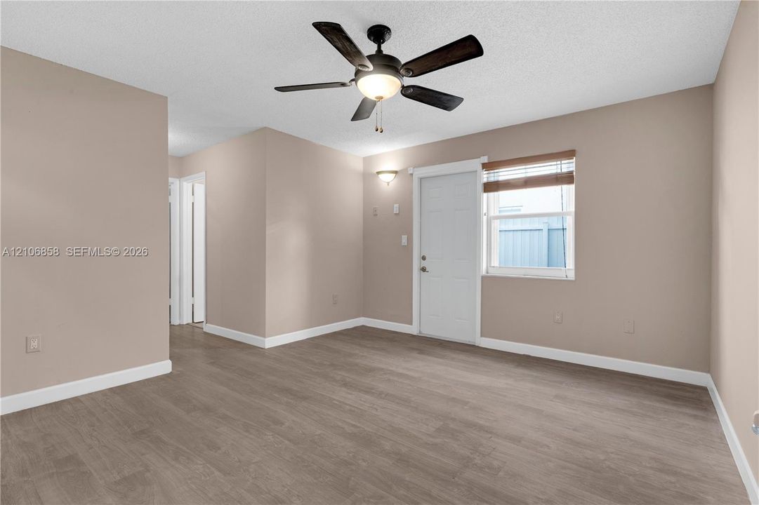 Neutral-toned room featuring wood-finish flooring, a central ceiling fan with integrated lighting, wall-mounted accent lighting, and a single-panel door