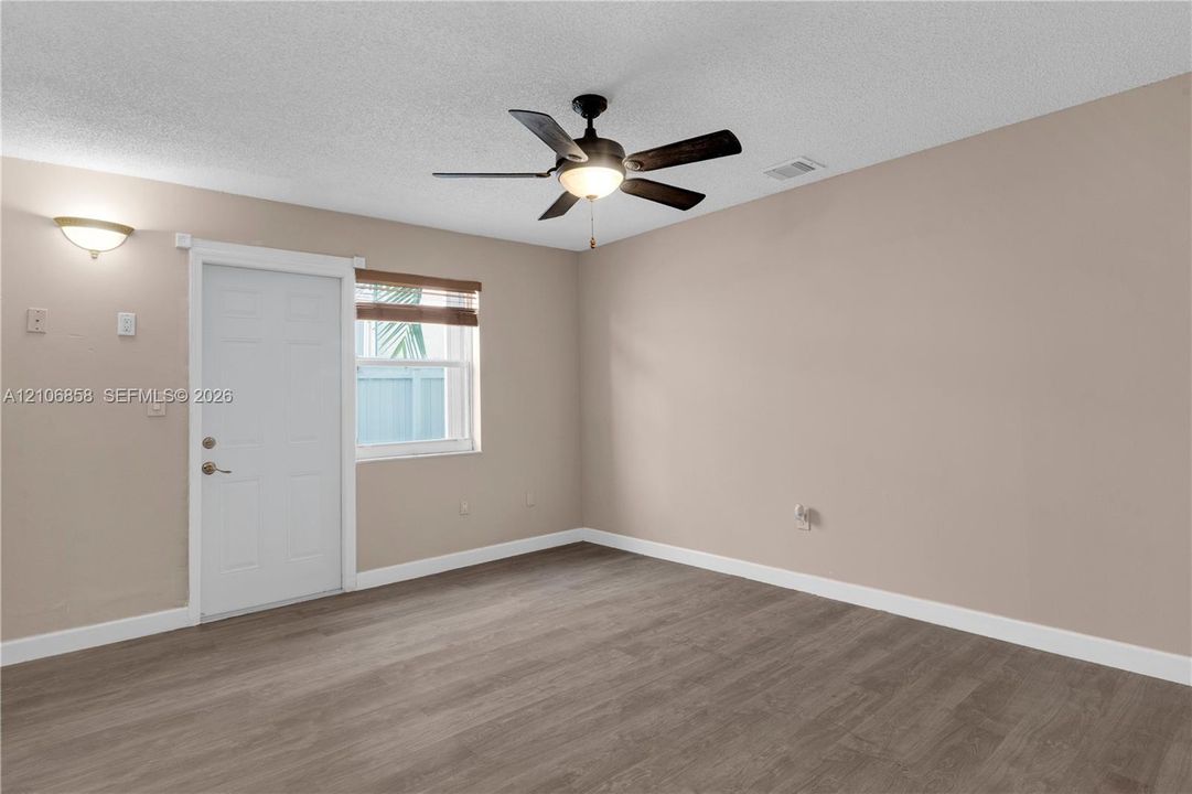 Neutral-toned interior room featuring wood-finish flooring, a ceiling fan with integrated lighting, a single exterior door, and a wall-mounted sconce