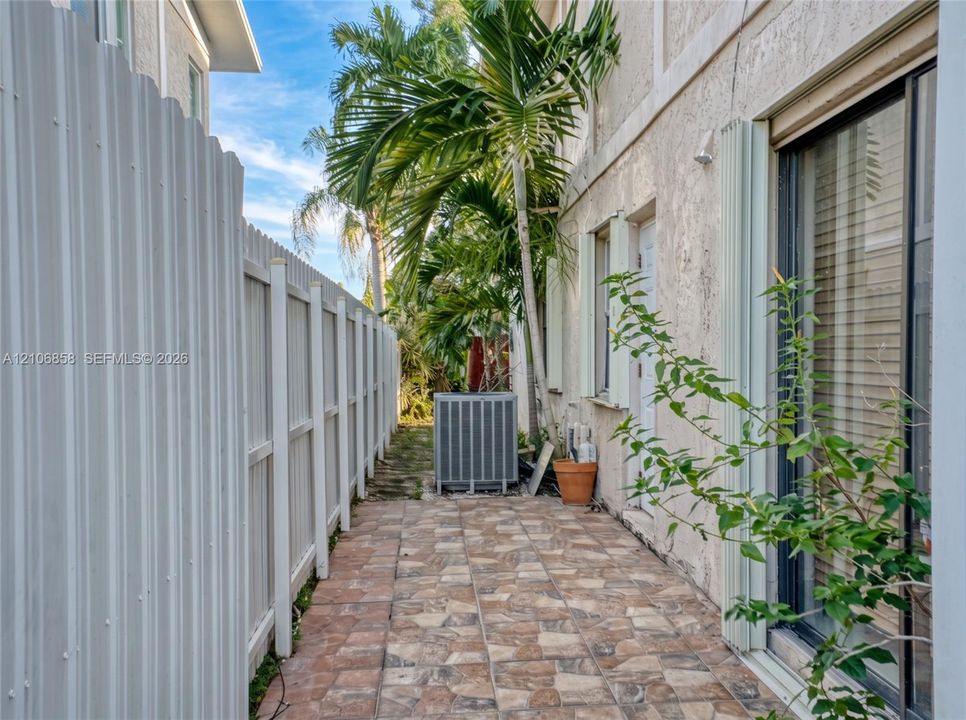 Side exterior featuring a paved stone-patterned walkway, white corrugated metal fencing, and stucco siding