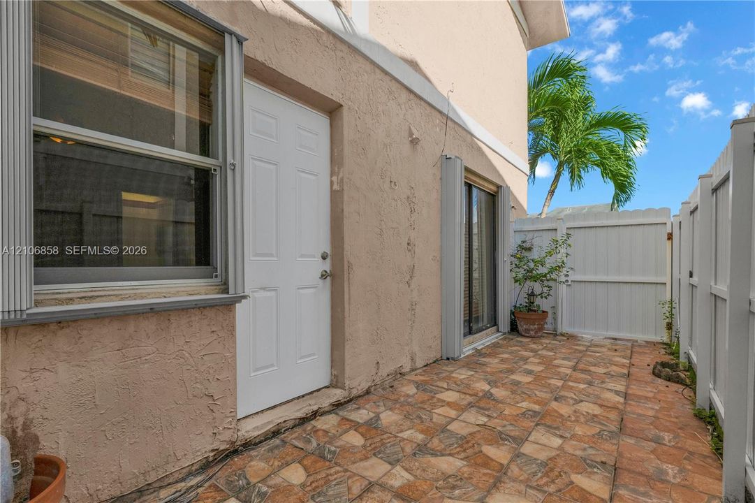 Stucco exterior with multi-pane window, white paneled entry door, sliding glass door, and tiled patio enclosed by white vinyl fencing