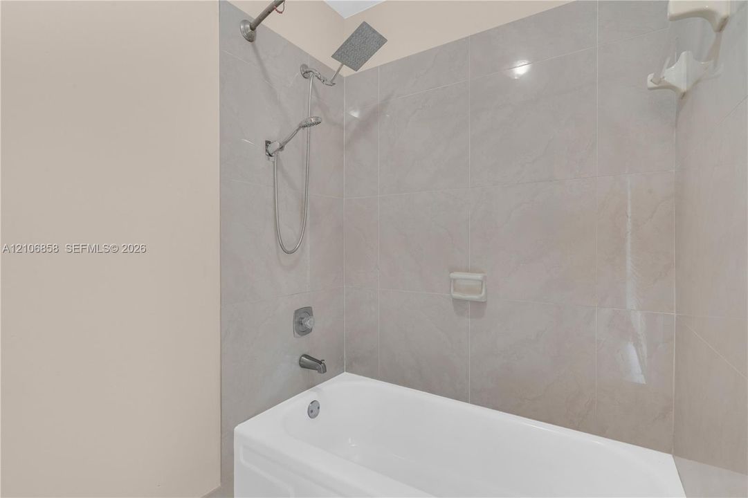 Bathtub surround with neutral-toned oversized wall tile, rainfall shower head, handheld shower fixture, and integrated soap niche
