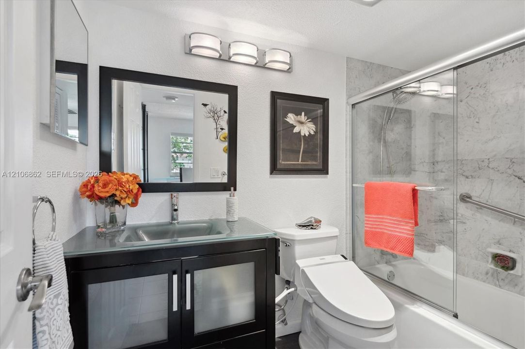 Bathroom featuring a vanity with a glass countertop and integrated sink, a deep-toned cabinet with frosted glass panel doors, a three-light wall-mounted fixture, and a glass-enclosed shower with stone-finish wall tile