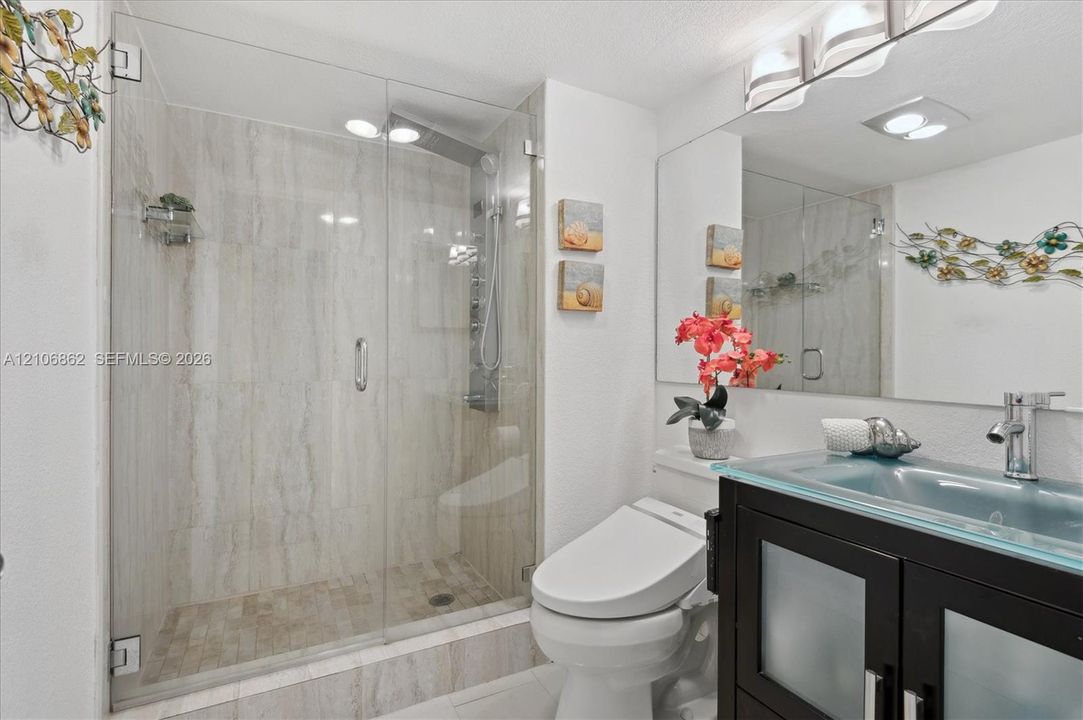 Walk-in shower with floor-to-ceiling stone-look tiling and frameless glass door, vanity with glass countertop and integrated sink, and dark wood-finish cabinetry with frosted glass inserts