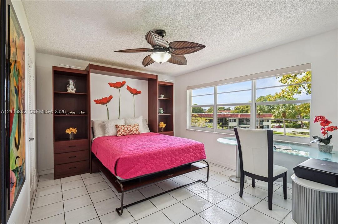 Built-in mahogany-tone cabinetry with integrated shelving and drawers, large multi-pane window, ceramic tile flooring, and ceiling fan with palm-leaf style blades