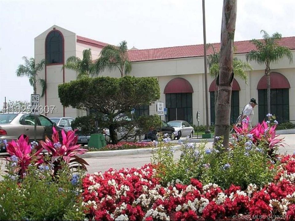 Commercial exterior featuring a light-toned stucco façade, red clay tile roof, arched window treatments, and landscaped grounds with mature palm trees
