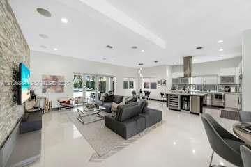 Open-concept living space featuring high ceilings with recessed lighting, large-format tile flooring, a floor-to-ceiling stone-veneer accent wall, and a kitchen equipped with a stainless steel range hood and built-in wine refrigeration