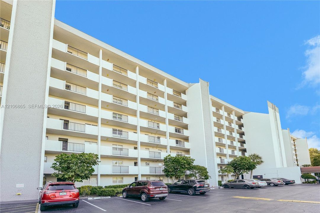 Multi-story residential building exterior featuring stacked private balconies with metal railings, a light-toned stucco façade, and an asphalt parking area