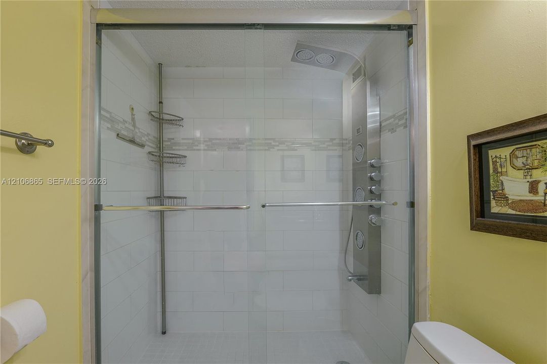 Shower enclosure featuring white tile walls, a horizontal glass and stone mosaic inlay, sliding glass doors, and a stainless steel multi-head shower panel system