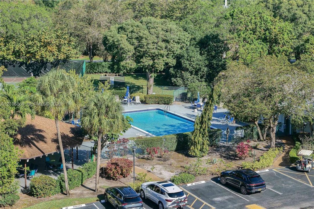 Rectangular outdoor swimming pool with surrounding concrete deck, covered pavilion, fenced tennis court, and mature trees