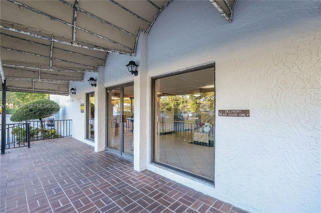 Exterior facade featuring a covered brick-paved walkway, stucco walls, large fixed glass storefront windows, and metal wall-mounted lanterns