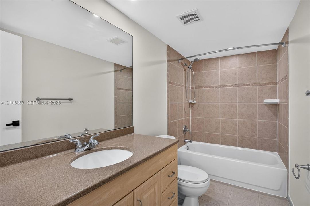 Bathroom featuring a light wood cabinetry vanity, solid-surface countertop with an integrated oval basin, full-width wall mirror, and a tub-shower combination with ceramic tile surround