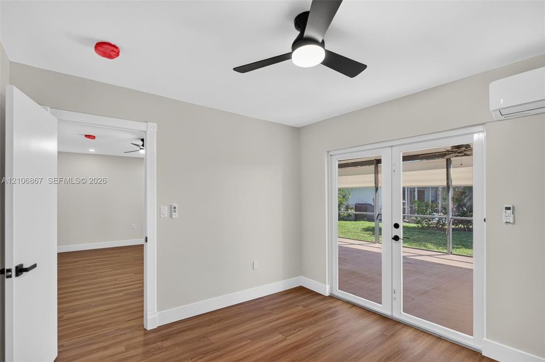 Wood-finish flooring, matte black ceiling fan with integrated lighting, wall-mounted ductless air conditioning unit, and double glass-paned doors opening to a covered patio