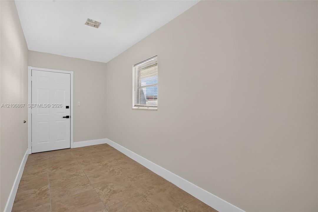 Neutral-toned interior space featuring light-colored wall paint, large-format tile flooring, a white paneled entry door with matte black hardware, and a single window with horizontal blinds