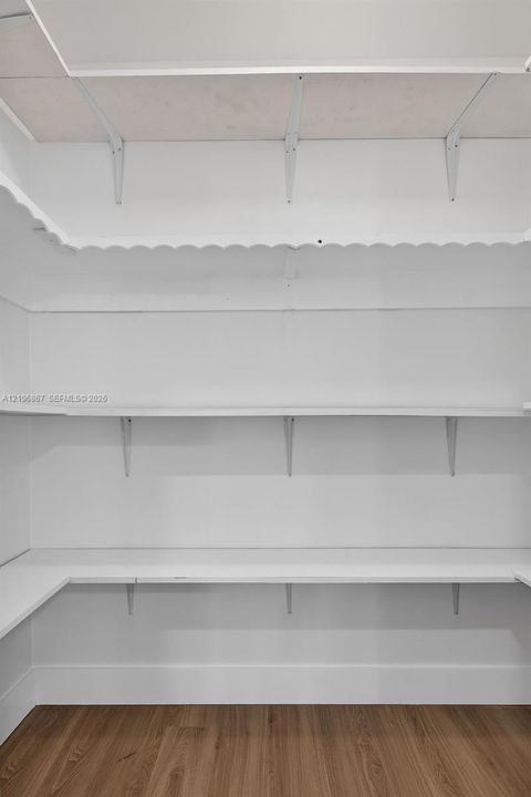 Walk-in storage space featuring multi-level white shelving with decorative scalloped trim, supported by sturdy wall-mounted brackets, finished with wood-finish flooring